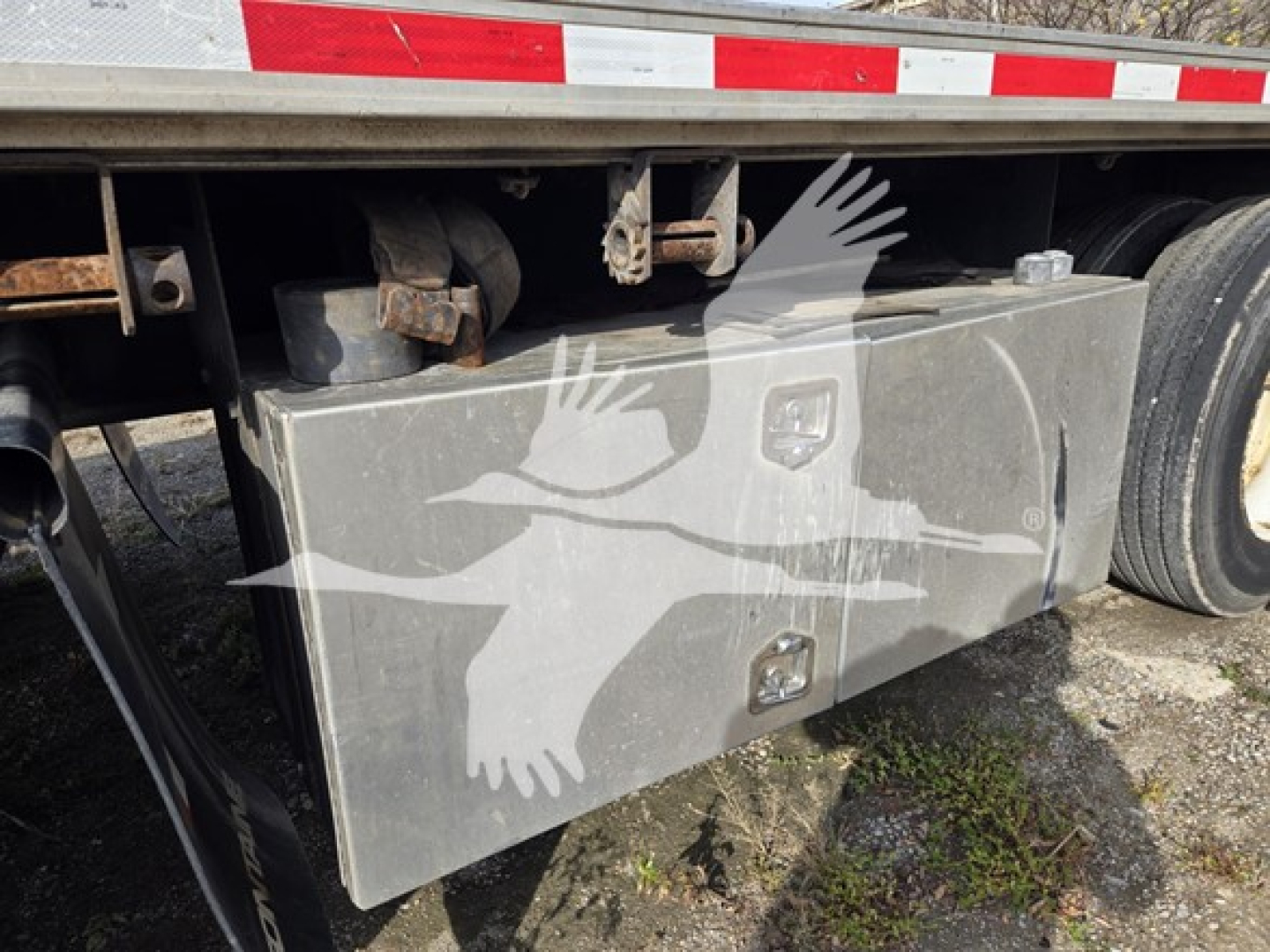Trailer side view showing a rectangular metal toolbox mounted under the flatbed of a Fontaine Infinity trailer with a dual wheel setup nearby.
