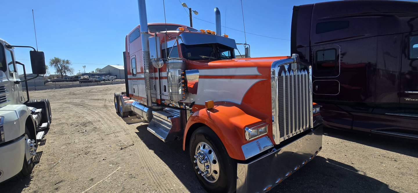 Kenworth W900 conventional sleeper truck in orange and white seen from front-left, chrome grille, large dual exhaust stacks.