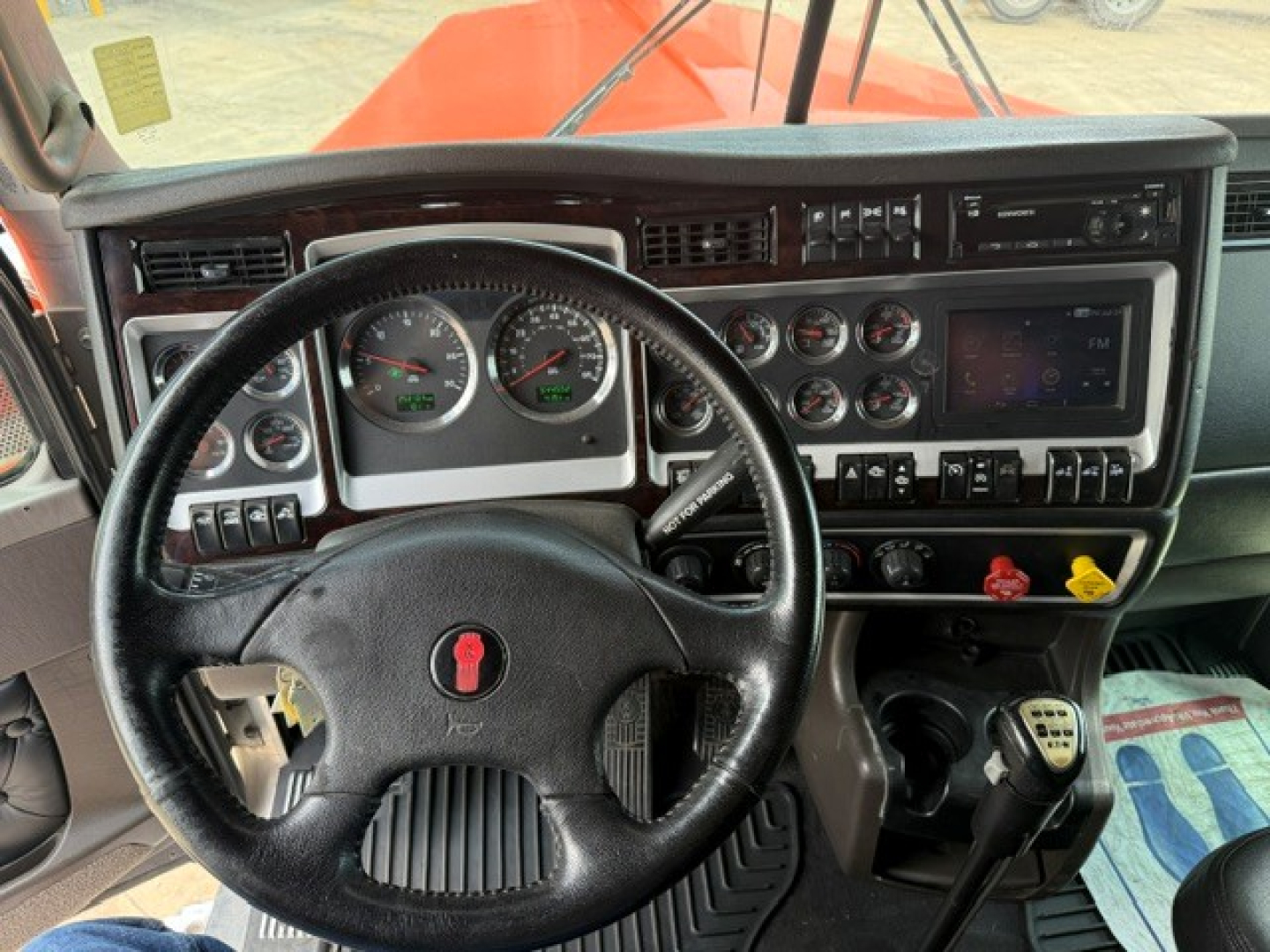 Kenworth W900 cab dashboard and steering wheel; multiple gauges, switches, and a touchscreen-like display visible. From the image: no explicit legible text other than general dashboard components.
