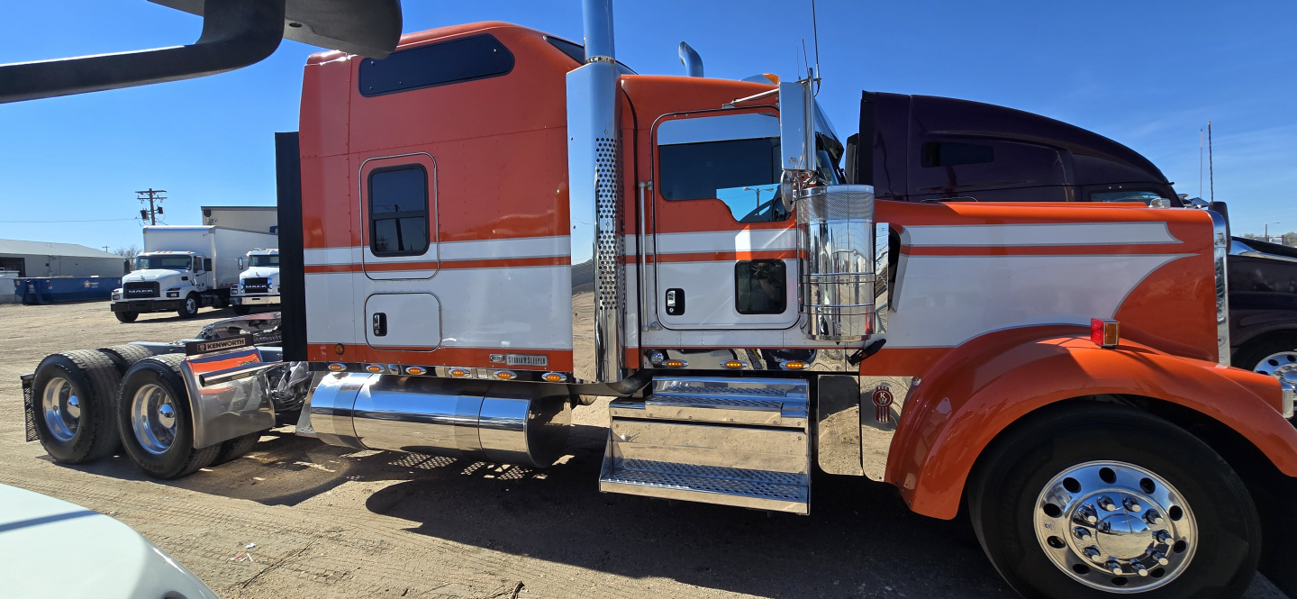 Kenworth W900 sleeper cab truck side view showing orange and white paint, chrome steps, fuel tanks, and dual rear axles.