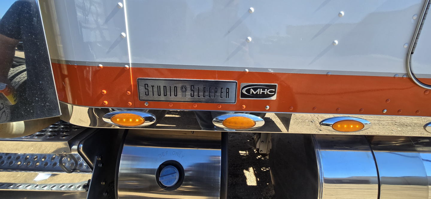 Kenworth W900 rear view showing Studio Sleeper badge and MHC logo on the tail with orange striping and chrome bumper.