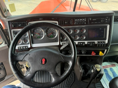 Kenworth W900 cab dashboard and steering wheel; multiple gauges, switches, and a touchscreen-like display visible. From the image: no explicit legible text other than general dashboard components.