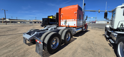 Kenworth W900 truck tractor in a dirt lot, orange sleeper cab, dual rear axles, chrome fuel tanks, viewed from rear-left side showing chassis and fifth wheel.