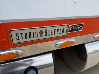 Studio Sleeper badge on Kenworth W900 truck showing MHC logo and chrome exhaust. Logo text: STUDIO SLEEPER with MHC badge; visible orange side marker lights.