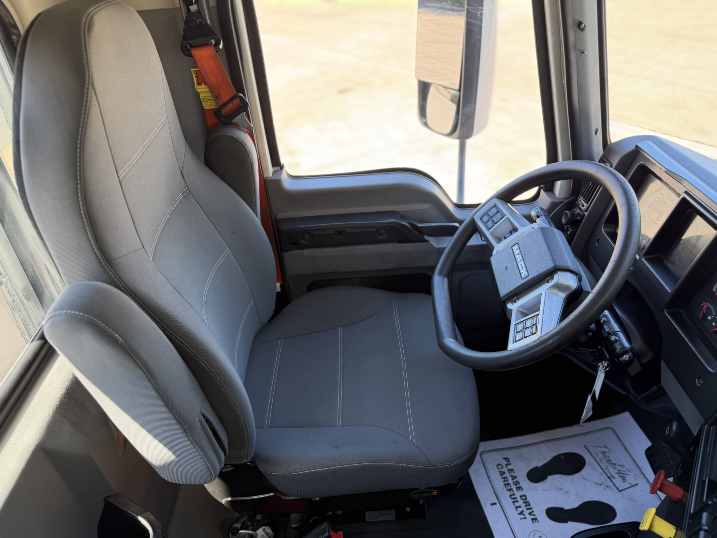 Truck cab interior showing steering wheel and driver's seat in a 2023 Mack ANTHEM 64T. Visible elements include gray cloth seat, dashboard, steering wheel with controls, and side window.