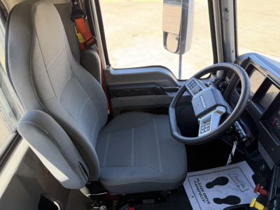 Truck cab interior showing steering wheel and driver's seat in a 2023 Mack ANTHEM 64T. Visible elements include gray cloth seat, dashboard, steering wheel with controls, and side window.