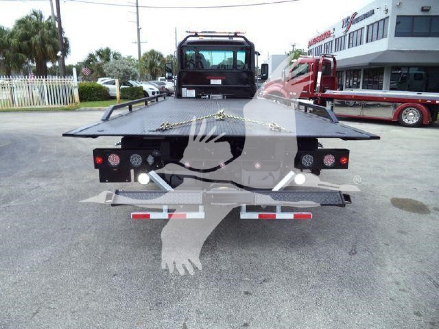 Flatbed rollback tow truck rear view showing platform and taillights with chrome bumper, Kenworth dealership background.