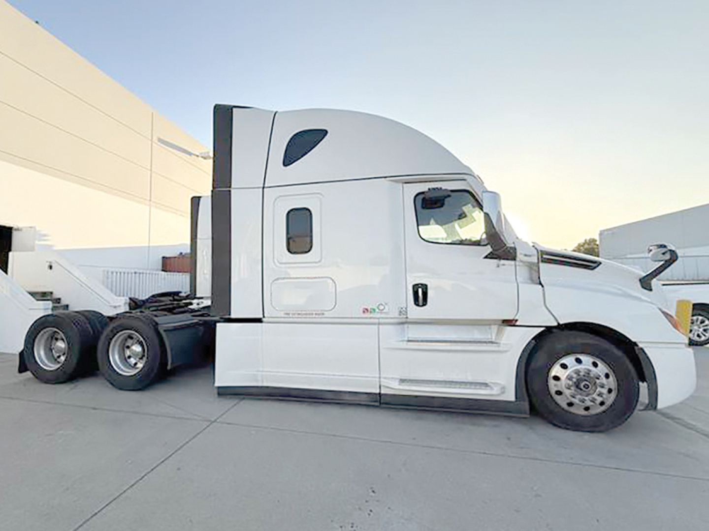 Truck side view showing Freightliner Cascadia sleeper cab with white paint, fuel tanks, and dual rear axles.