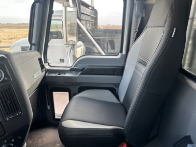 Cab interior of a 2026 Mack GR64F dump truck showing gray fabric seats, dashboard, and door panel; window reveals exterior heavy equipment.
