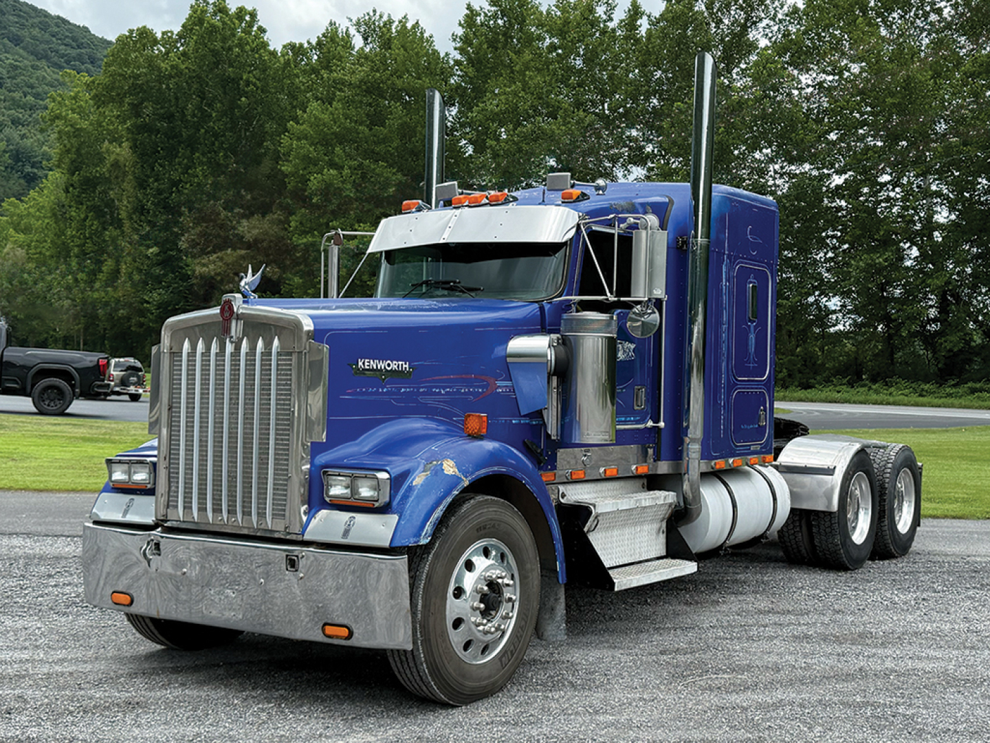 Kenworth W900L AeroCab sleeper truck in blue, front three-quarter view showing chrome grille, dual exhaust stacks, and large fuel tanks.