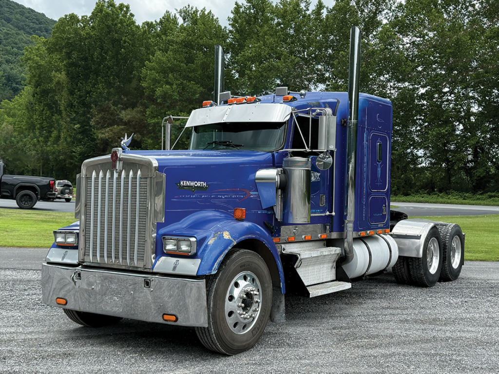 Kenworth W900L AeroCab sleeper truck in blue, front three-quarter view showing chrome grille, dual exhaust stacks, and large fuel tanks.