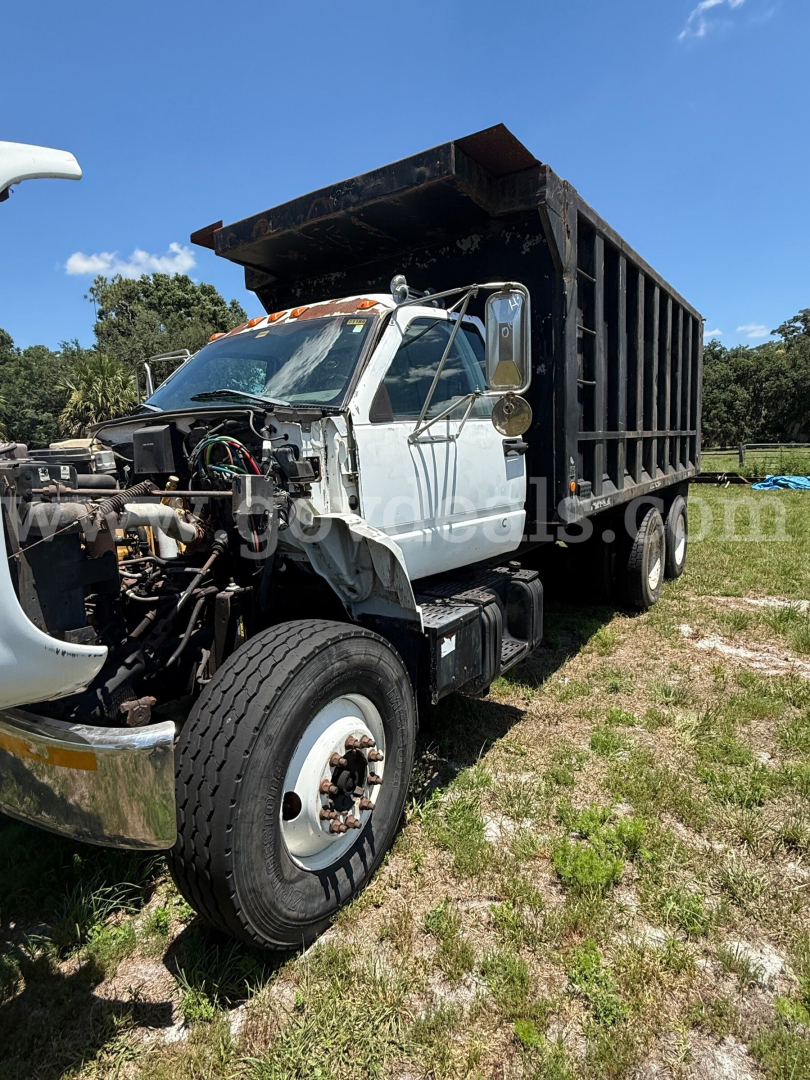 GMC dump truck with cab front removed showing engine bay and exposed components, silver cab, black dump bed, large front wheel, working site grass field