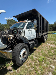 GMC dump truck with cab front removed showing engine bay and exposed components, silver cab, black dump bed, large front wheel, working site grass field