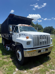 GMC dump truck showing front grille and side profile with dump bed raised, white cab, chrome bumper, and orange warning lights.
