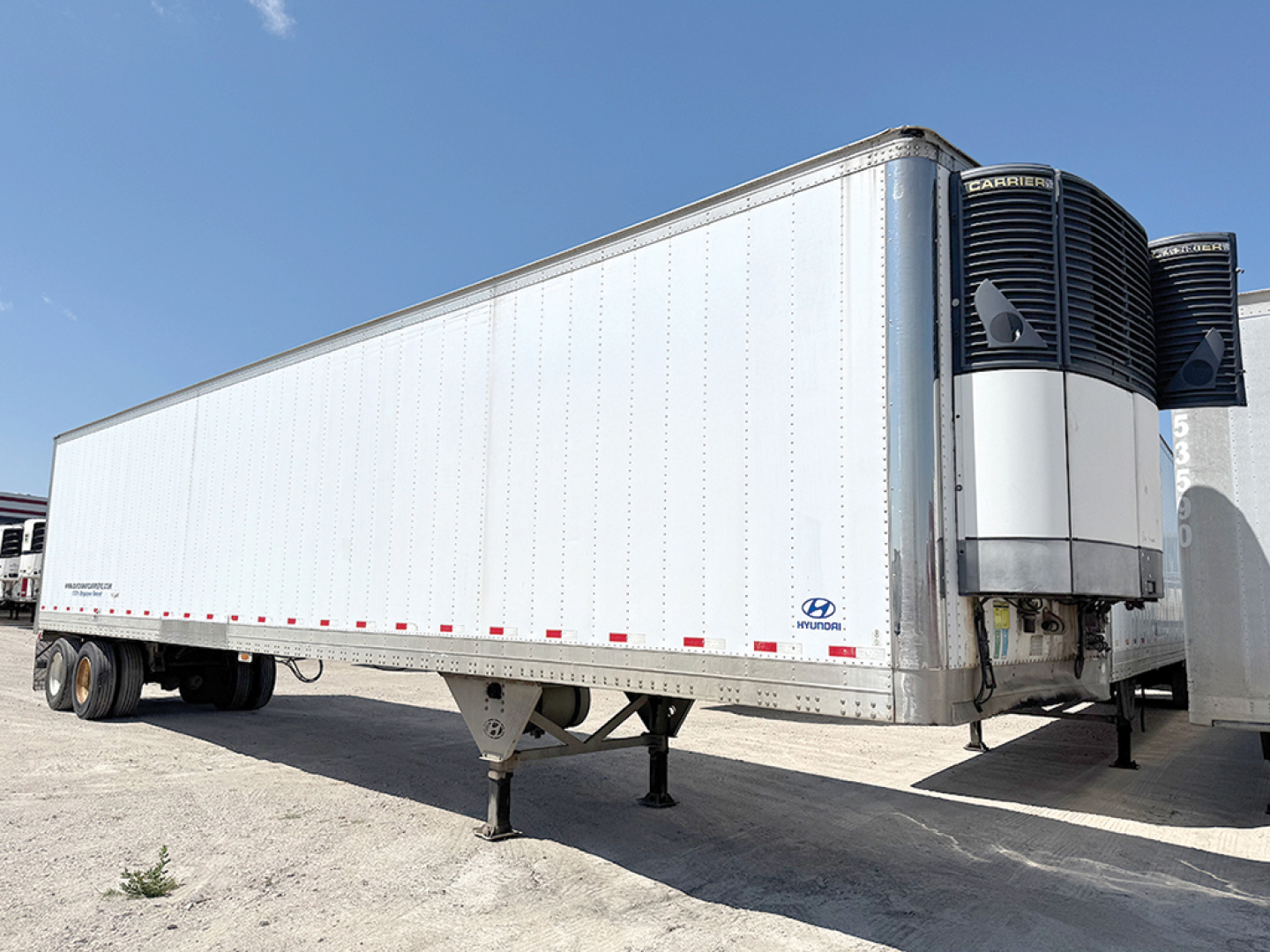 Hyundai aluminum reefer trailer with front refrigeration unit and tandem rear axles, side view, clean metal walls, blue sky.