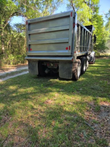 Rear view of a dump truck with a metal dump bed, dual rear wheels, and mud flaps. The trailer shows tailgate panels and taillights.
