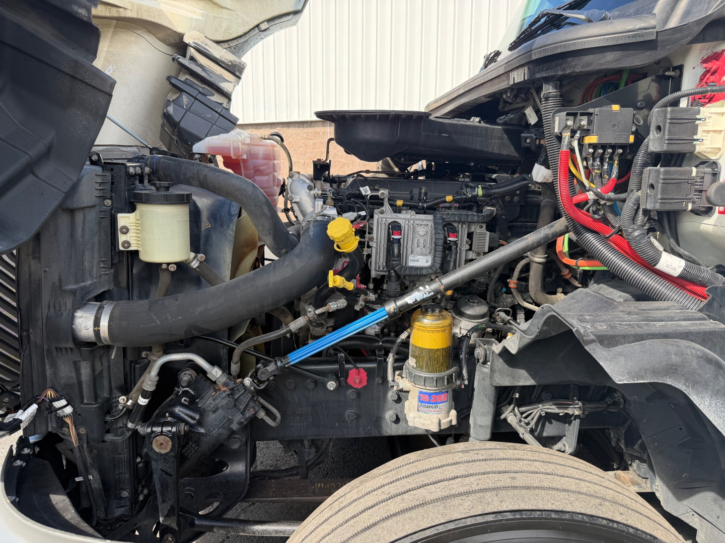 Truck engine bay of a used 2021 International LT625 with visible coolant reservoir, air filters, hoses, wiring, and yellow dip stick; front wheel and suspension components visible.
