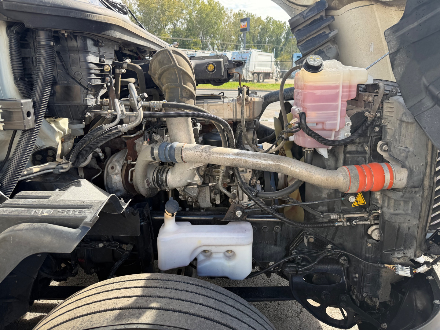 Truck engine bay of 2021 International LT625 showing coolant reservoir, pink/red coolant expansion tank, white windshield washer reservoir, various hoses and metal lines.