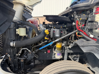 Truck engine bay of a used 2021 International LT625 with visible coolant reservoir, air filters, hoses, wiring, and yellow dip stick; front wheel and suspension components visible.