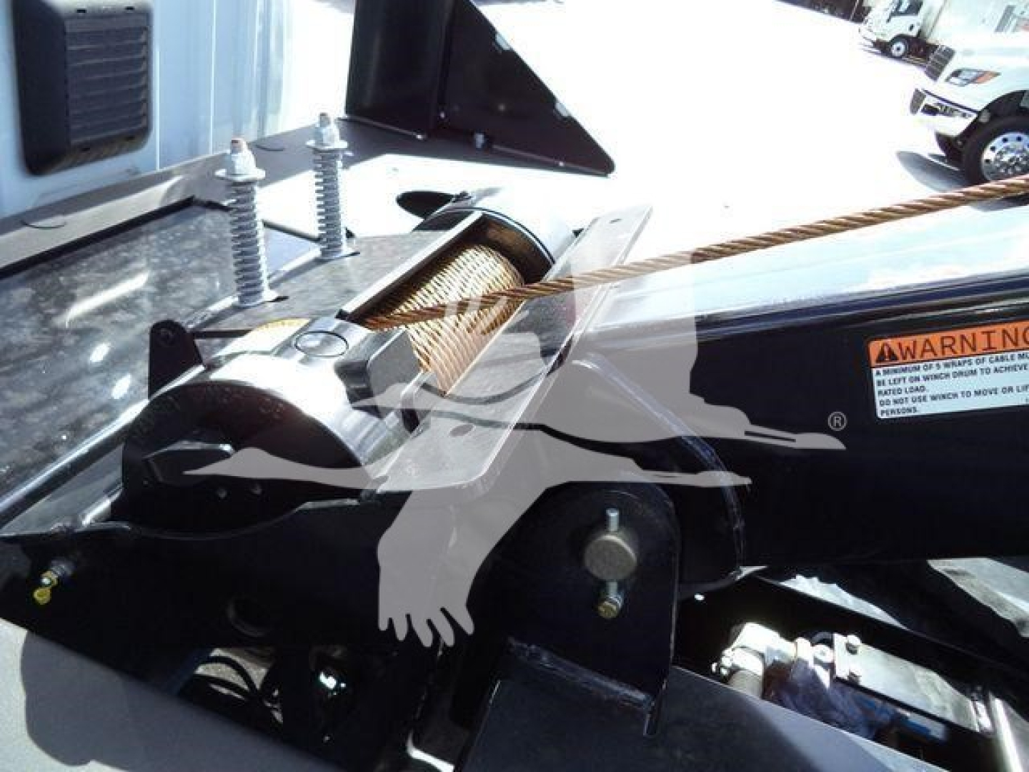 Truck bed area showing a winch mounting and hydraulic components with warning label and a roller fairlead. Visible text includes a small orange warning label and some metal fittings.