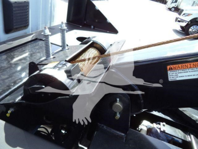 Truck bed area showing a winch mounting and hydraulic components with warning label and a roller fairlead. Visible text includes a small orange warning label and some metal fittings.