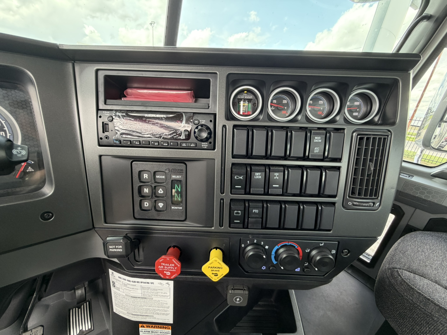 Dashboard view of a new Mack GR64B truck showing instrument cluster, switches, and control knobs in the cab. visible text includes gauges and yellow and red knobs.