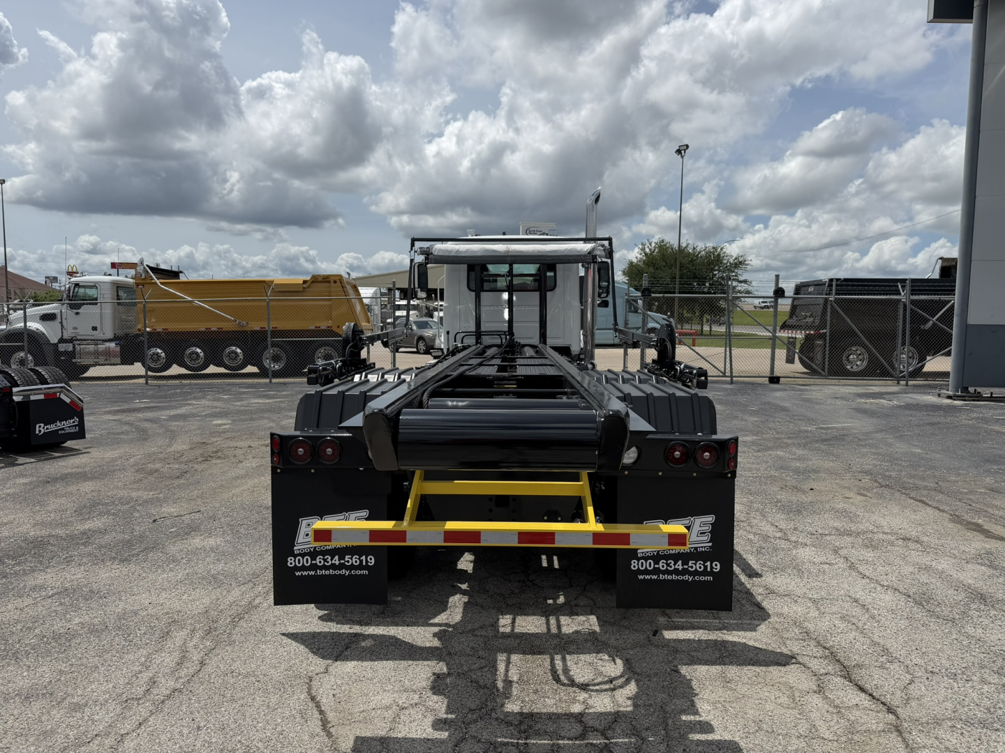 Rear view of a new 2026 Mack GR64B roll-off truck with trailer hookup, black chassis, and yellow safety stripe. Brand decals and contact numbers visible on mud flaps.