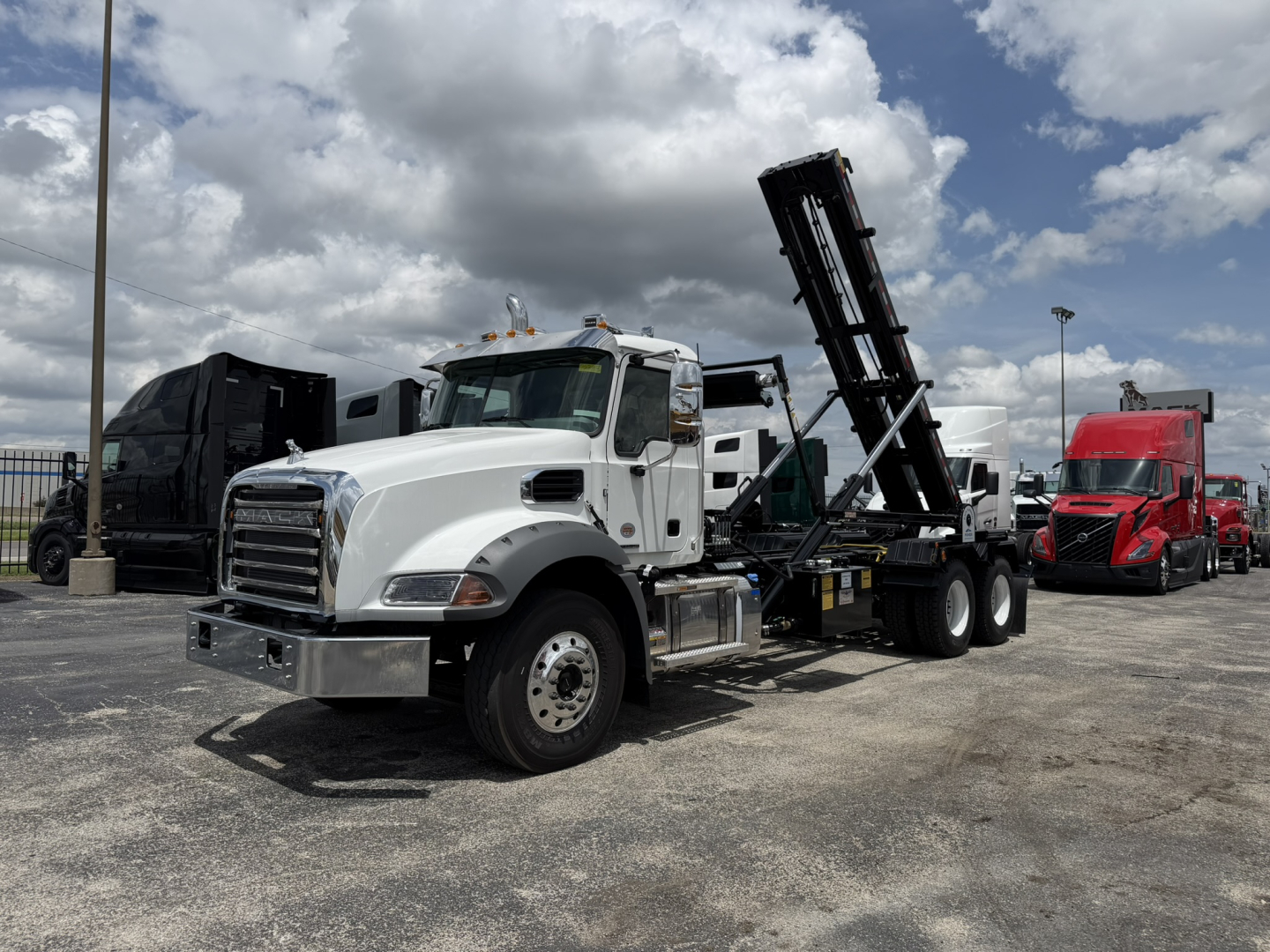White Mack GR64B roll-off truck in a parking lot, cab with chrome bumper and a raised hydraulic bed lift, other trucks visible in background.