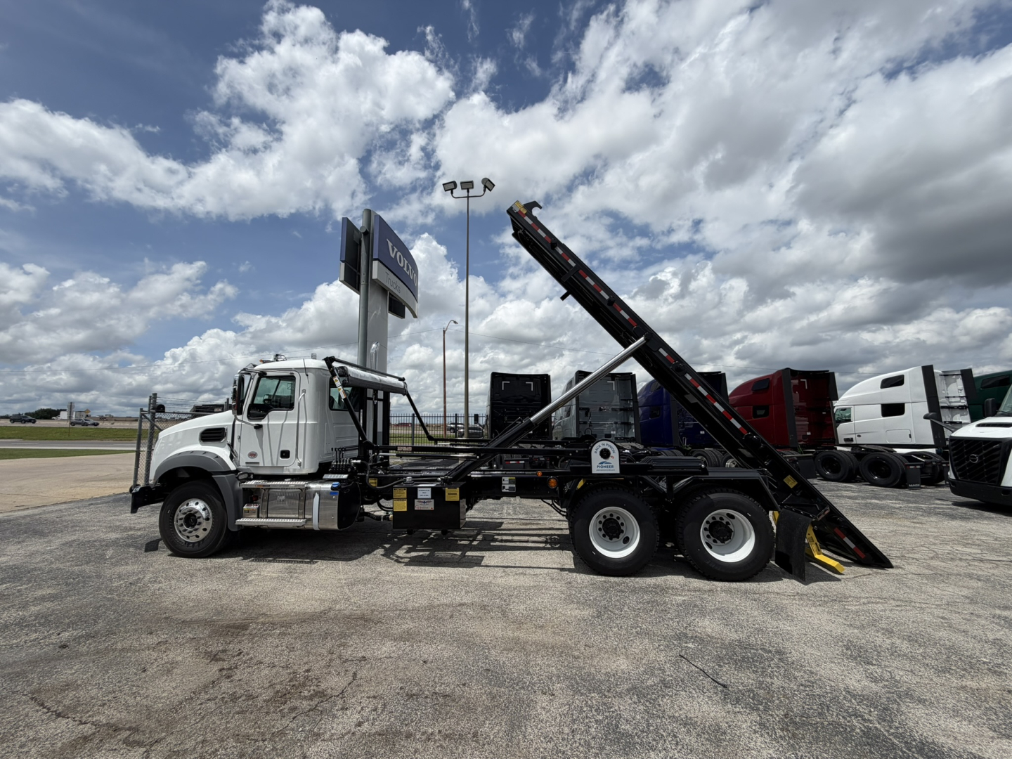 Mack GR64B roll-off truck side view showing hydraulic riser and rear loading ramp with black conveyor/ladder, white cab, multiple axles, in a dealership lot.