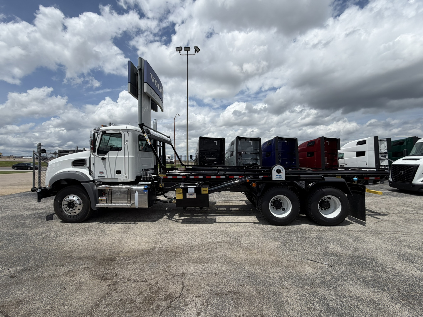 Mack GR64B roll-off truck in profile view showing dump bed and multiple cabs in background.