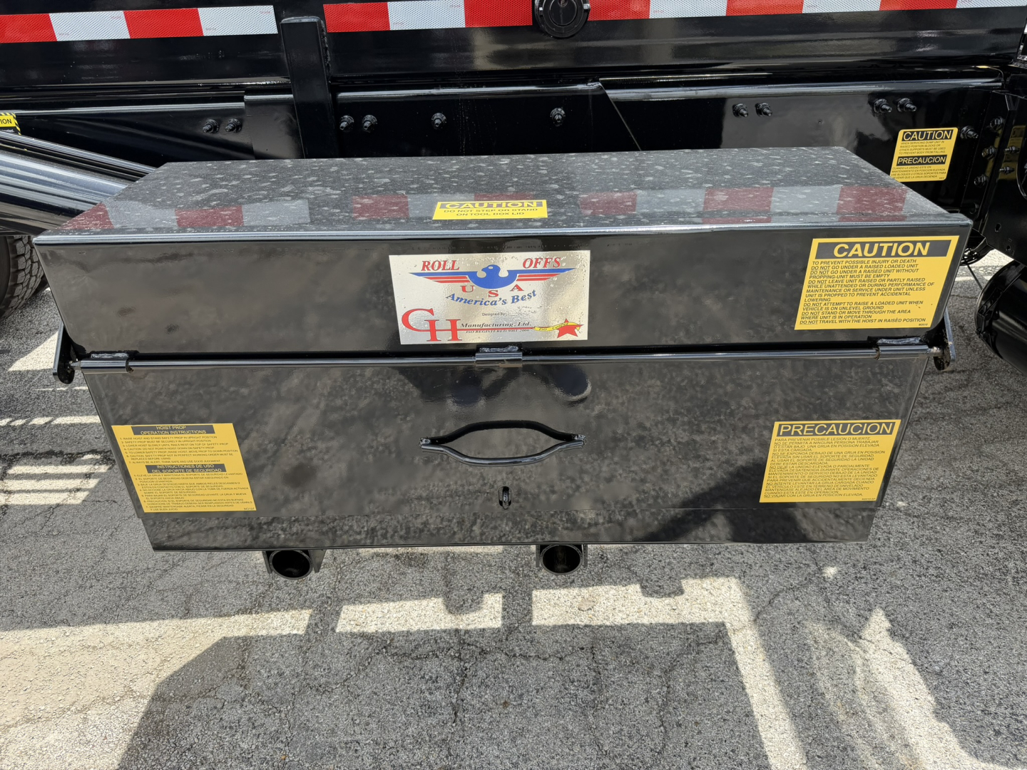 Rolloff truck toolbox mounted on Mack GR64B chassis showing multiple yellow caution labels and a central handle with a lock. Brand label reads Roll Offs Aluminum Toolbox.
