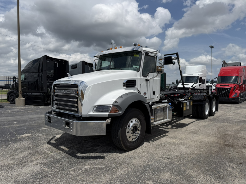 White Mack GR64B truck in a yard showing front and side, with a roll-off body and chrome bumper; chrome mirrors and steps visible; other trucks in background.