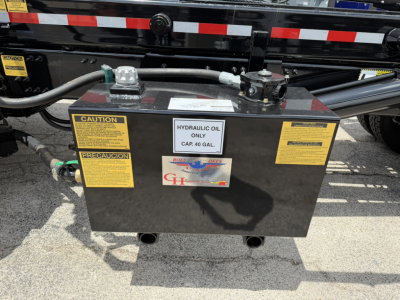 Rectangular hydraulic oil reservoir with yellow caution labels and a central white label reading HYDRAULIC OIL ONLY CAP 40 GAL. Black metal tank with hoses connection and a small sticker showing a logo.”