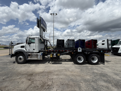 Mack GR64B roll-off truck in profile view showing dump bed and multiple cabs in background.