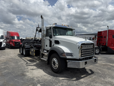 Tow truck/rollback truck in a lot with a white cab and flatbed, chrome bumper, front grille, and dual rear axles; Mack GR64B branding not clearly visible; clear blue sky with clouds.