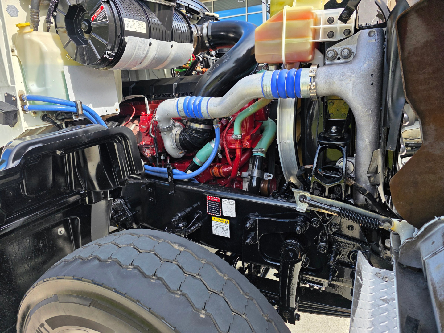 High-detail view of a Mack GRANITE truck engine bay showing coolant reservoir, hoses, radiator, and engine block with red components and blue clamps.