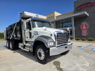 Mack Granite dump truck in front of dealership building. Vehicle shows front grille, chrome bumper, dual exhaust stack, aluminum flatbed with dump body, and branding; visible text includes MACK on grille and KW logo on building.