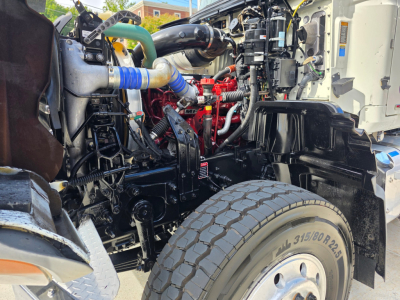 Engine bay of a 2022 Mack GRANITE dump truck showing exposed diesel engine with red components, blue silicone hose, green coolant hose, and wiring harnesses.