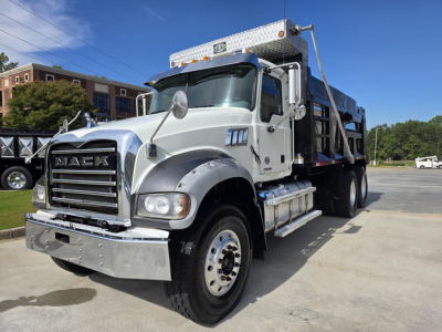 Mack Granite dump truck front left side with chrome grille, large bumper, dual exhaust stack, and dump bed in background.