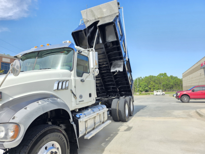 Dump truck tipping with raised cargo bed on Mack Granite chassis, white cab, multiple stacked wheels, VIN visible in listing; side mirrors and hydraulic lines present.