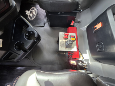 Truck interior showing center console with controls and a red switch panel adjacent to a cubby; floor mat with Mack logo visible.