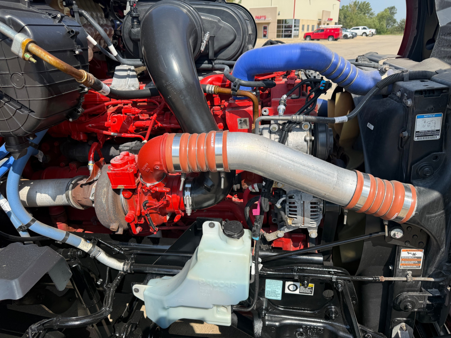 Kenworth T680 engine bay showing red engine, large silver intake pipe, blue silicone hoses, black radiator with coolant reservoir, and various hoses and wiring.