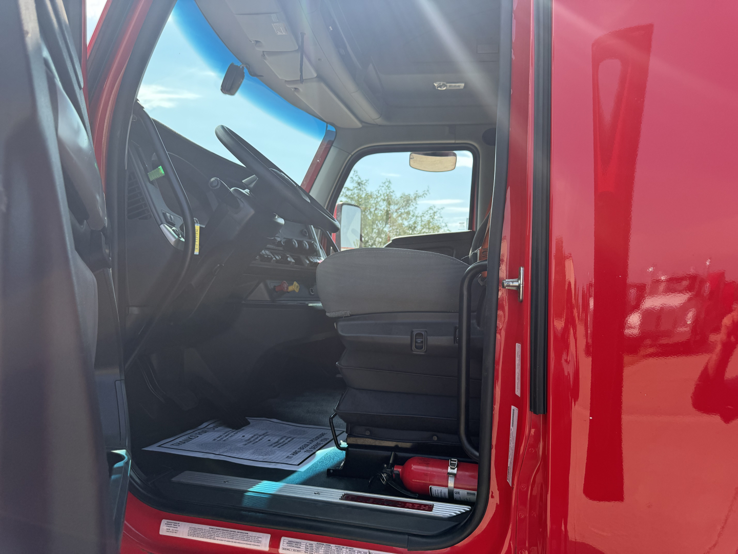 Interior of a Kenworth T680 cab showing driver seat, steering wheel, and dashboard; red exterior door frame; floor mat and a small red extinguisher visible under the seat.