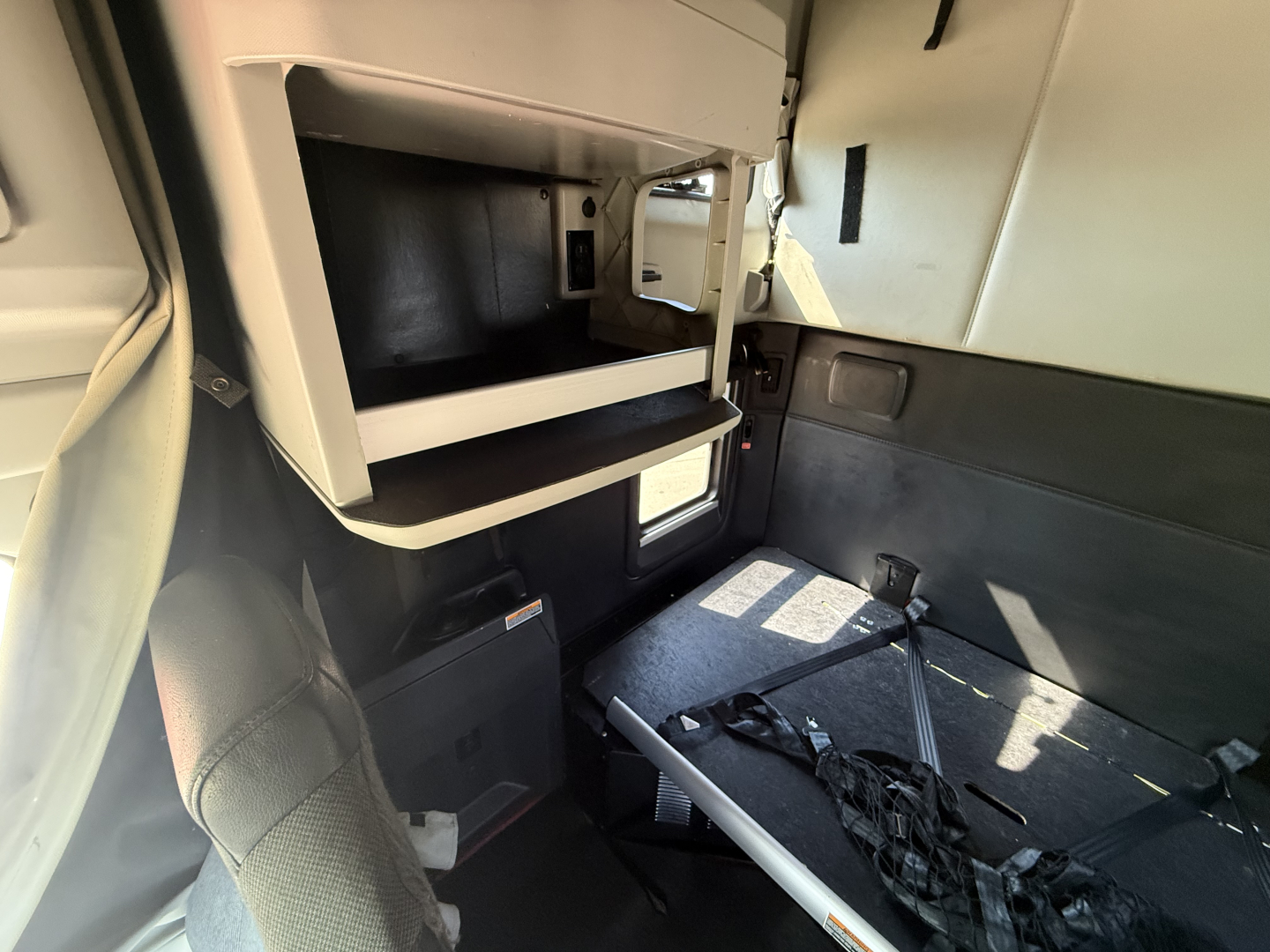 Interior of a used 2024 Kenworth T680 sleeper cab showing storage shelf and open compartments with seating area and seat belts.