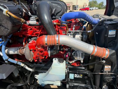 Kenworth T680 engine bay showing red engine, large silver intake pipe, blue silicone hoses, black radiator with coolant reservoir, and various hoses and wiring.