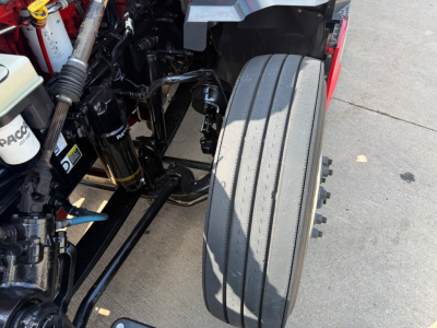Kenworth T680 front wheel and engine bay view showing steering components and suspension; visible components include air filter housing, hoses, and chassis frame.