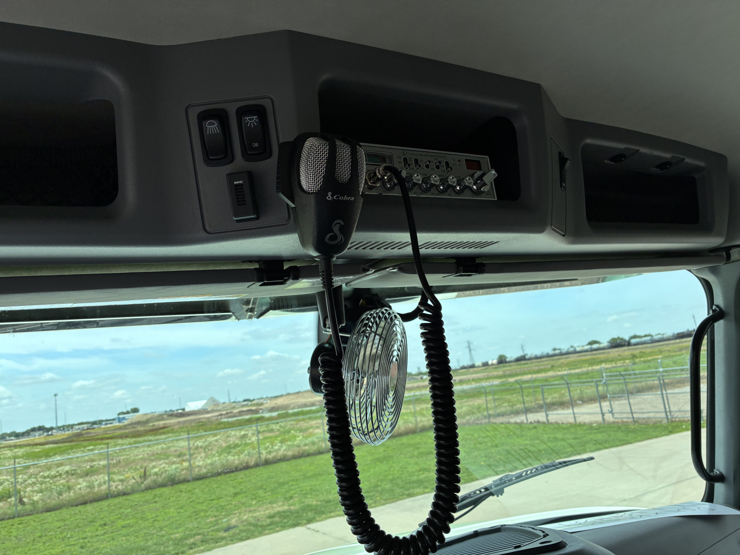 Truck cab interior showing CB radio mic and control panel mounted over windshield with vents and switches in view; exterior field visible through front window.