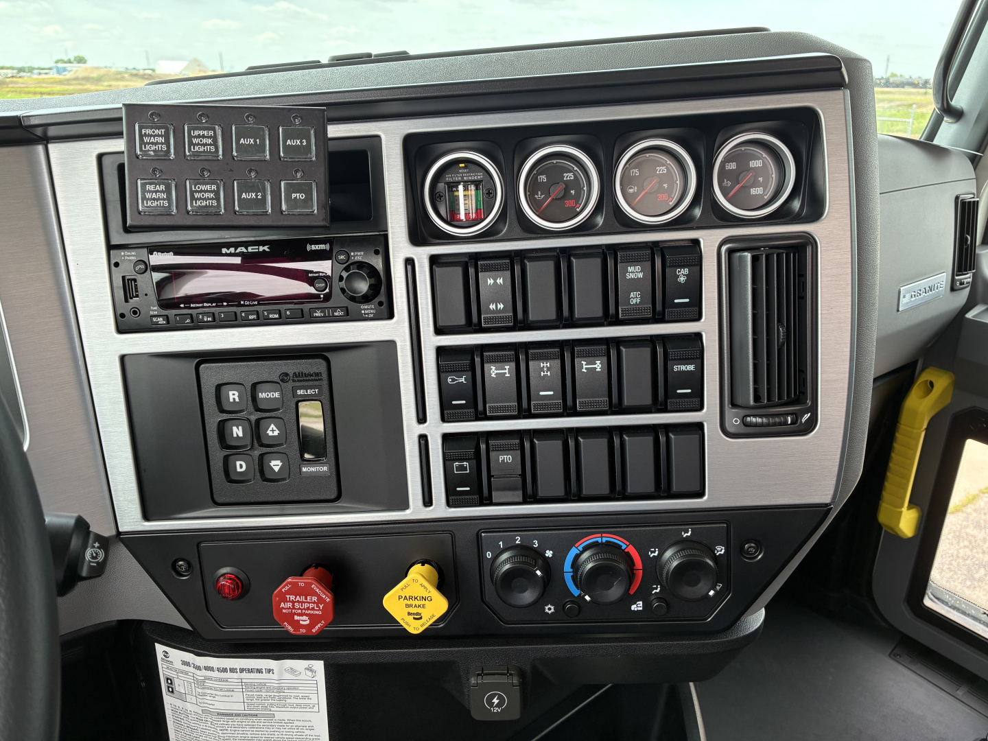 Mack GR64F dashboard showing multiple gauges, climate controls, radio, buttons, and switches in a truck cab interior.