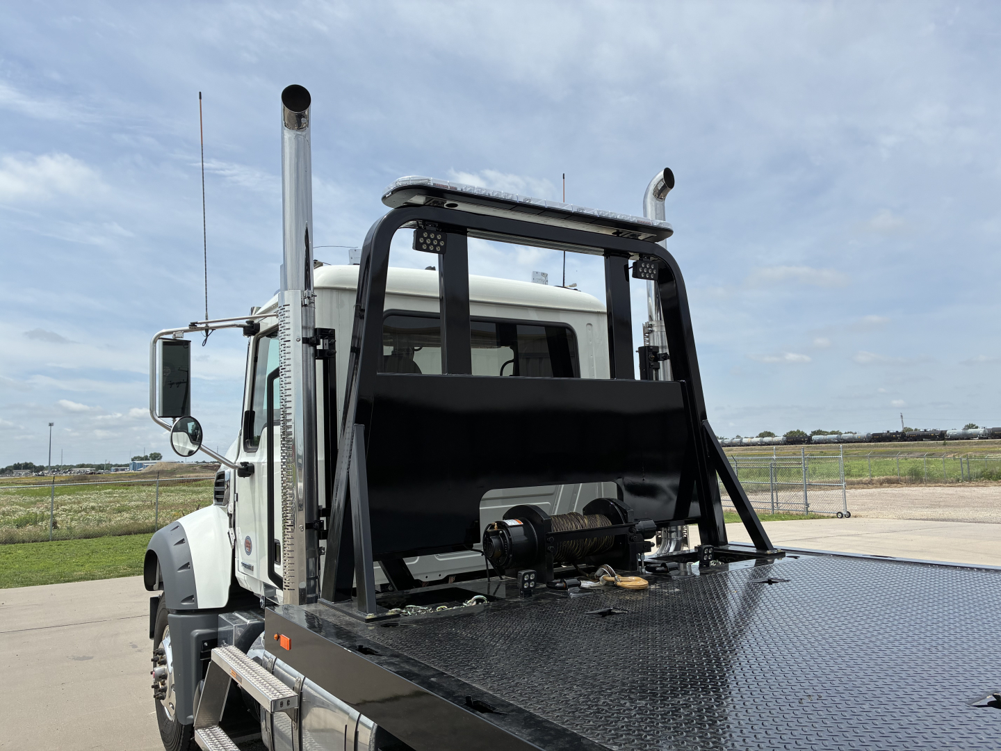 Truck cab with a metal flatbed and roll-back winch apparatus, tall exhaust stacks, and protective rollover bars. Light-colored truck in open lot.