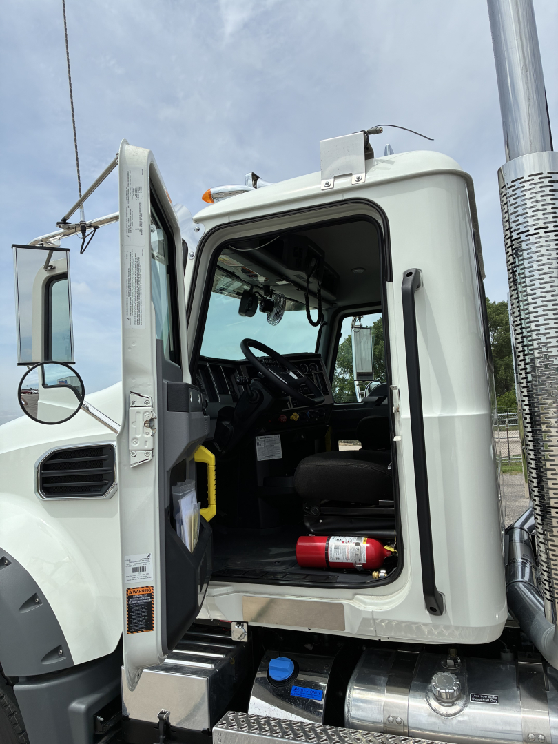 Truck cab interior visible through open door of a white Mack GR64F sleeper. Steering wheel, driver's seat, and control panels visible; blue bottle and red fire extinguisher in the step area.